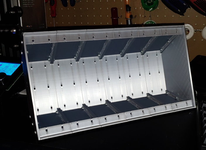 An empty silver box, the top and bottom taper towards the back. The inside is lined with ribs with bolts through them.