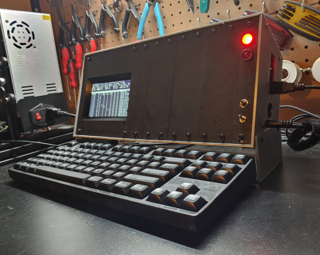 A silver box with a black face. A computer screen, LEDs and switches are set into the face. A keyboard sits in front of it.