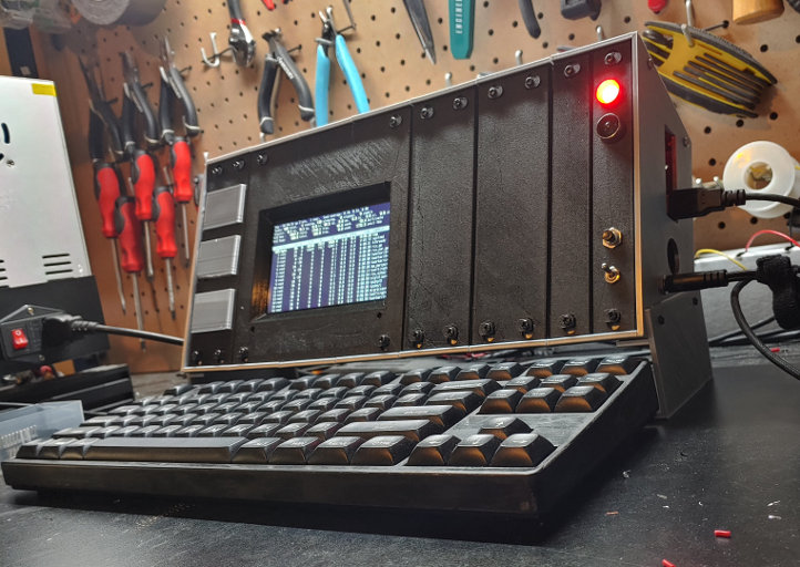 A silver box with a black face. Silver panels, a computer screen, LEDs and switches are set into the face. A keyboard sits in front of it.
