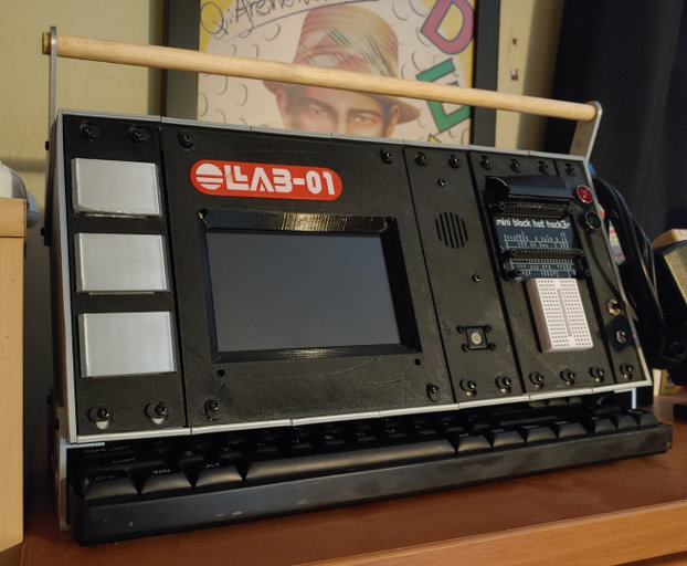 A silver box with a black face. Silver panels, a computer screen, LEDs and switches are set into the face. A keyboard is tucked under it.