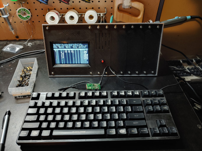 A silver box with a black face. There is a screen in the left side of the face. A red button and wires come out of the bottom middle of the face. The right side is featureless black.