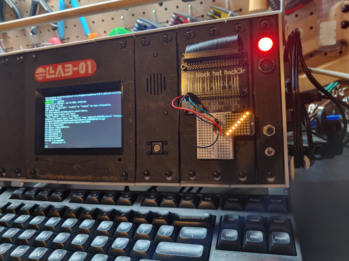 The black panels have been modified. One has a speaker grill and a tiny white ball mounted in it. Another has a ribbon cable coming out of it and plugging into a black PCB with multiple headers on it. Below the PCB is a small breadboard, with jumpers connected to the PCB headers, and a LED matrix plugged into it.