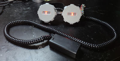 Round silver goggles. Where the lenses would be, there are silver cylinders with gear-like protrusions around the edges and only small rectangular openings. In each openings is 10 rectangular LED segments in a row. A black cable runs above the bridge, and a circuit board wires hang out of the eyepiece on the right (wearer's left).