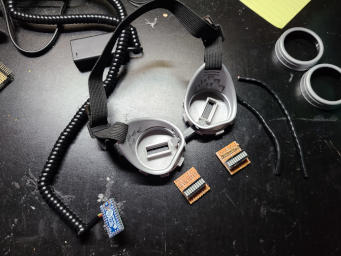 The parts laid out before wiring. There are two circuit boards with LED modules and resistors, a coiled cable with a battery box at one end and a circuit board at the other, some lengths of wire, the discarded original lens holders, and the modified goggles plastic.