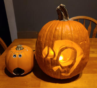 A large pumpkin with an image of a cartoon robot with a comical expression carved into it. A smaller pumpkin next to it has two metal eyes and round speaker in the place of a mouth.