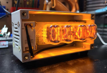 The circuit boards and a beefier fan, mounted in a silver box. The front of the box is open. The bottom stands elevated on black plastic feet, barely visible in shadow. There is a grill and screws on the side holding the fan.