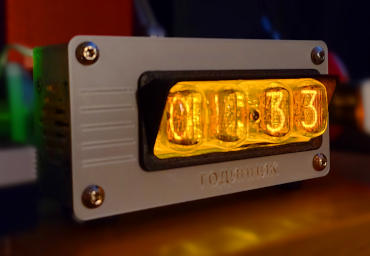 An oblong silver box standing on black feet. Four glass tubes containing neon-orange digits extend out of an opening in the front. They are framed by a black piece that projects over the top edge of them, like an awning. The front of the box has large button-head screws in the four corners, five inset lines decorating a space to the left of the tubes, and the word 'hodynnyk' in Ukrainian Cyrillic written below the tubes.