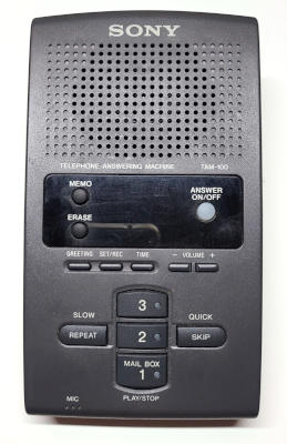 A charcoal-gray plastic slab. Most of the features are on the top surface. A Sony logo and speaker at one end. A display under clear plastic in the middle, with some buttons embedded in the lens. Small lozenge shaped buttons past that, then larger buttons on the other end.