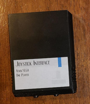 A black plastic cartridge labeled 'Joystick Interface: Atari/Sega'. A pair of small screws protrude from the end.