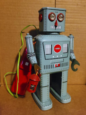 A boxy humanoid robot. Two domed lamps form its eyes, a sharp nose projects from its face, and it has a narrow vent for a mouth. The robot carries a lantern in its right hand. A wire runs from the robot's back to an oblong box with a button on it.