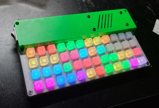 A rectangular plastic slab. There is a raised green section along the back with a rocker switch and a speaker grill. The lower section is silver with a 12x4 grid of light-up square buttons.