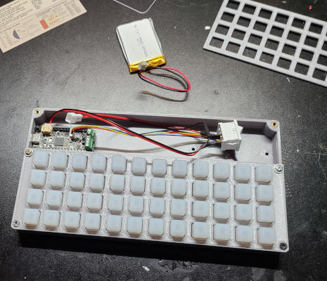 A silver plastic box containing a circuit board, a grid of buttons, wires, and a switch. A small Lithium Polymer battery is nearby.